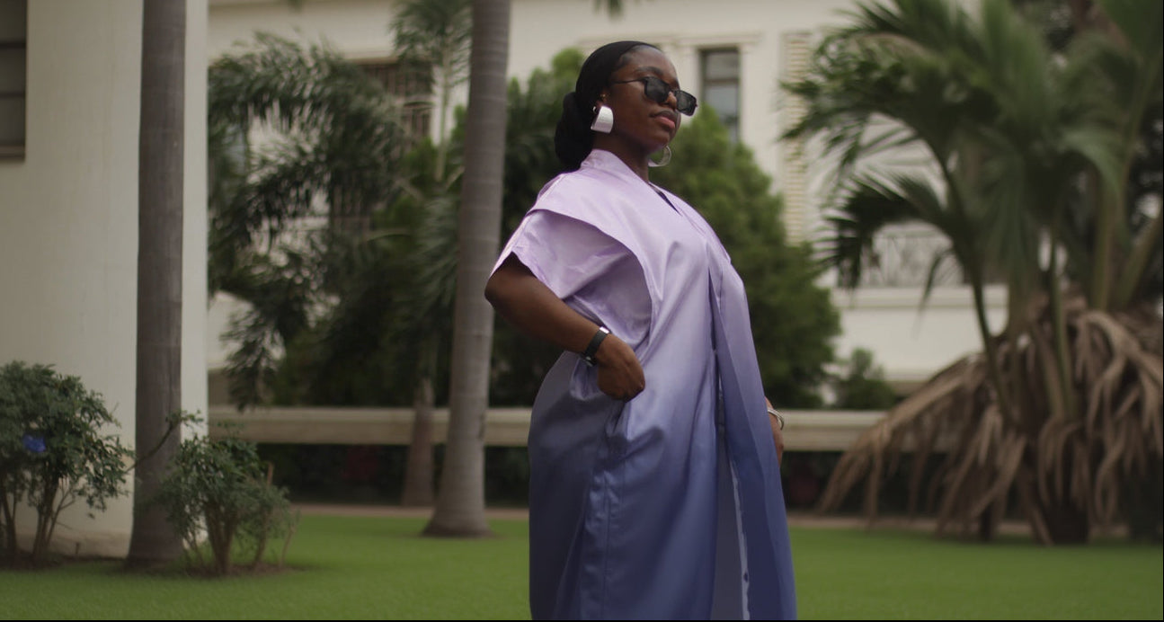 Woman in a light purple outfit walking outdoors with palm trees and a building in the backgroun